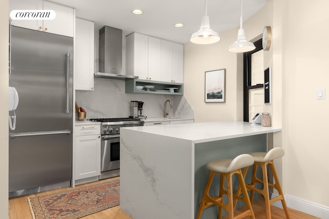 kitchen with high end appliances, white cabinets, wall chimney range hood, light wood-type flooring, and kitchen peninsula