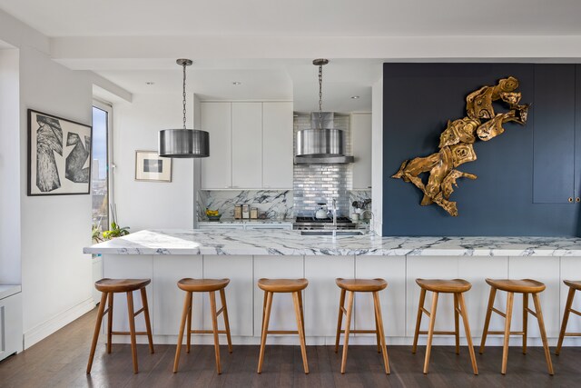 kitchen featuring pendant lighting, a kitchen breakfast bar, wall chimney exhaust hood, dark hardwood / wood-style flooring, and white cabinetry