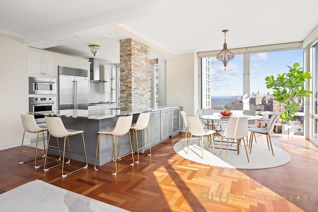 kitchen featuring white cabinets, wall chimney range hood, appliances with stainless steel finishes, decorative light fixtures, and a breakfast bar area