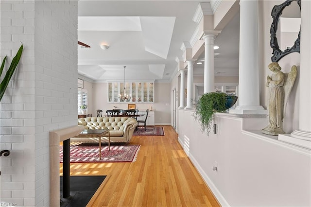 corridor featuring brick wall, a raised ceiling, light hardwood / wood-style floors, ornate columns, and an inviting chandelier