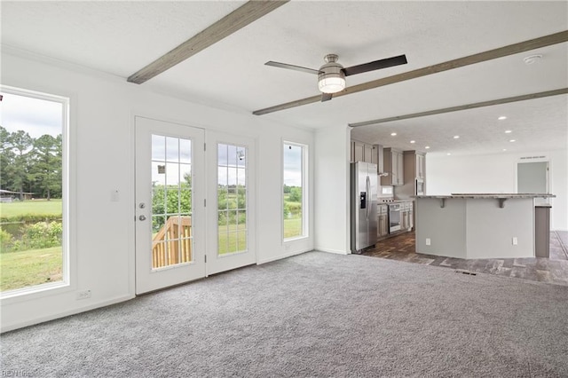 unfurnished living room with plenty of natural light, dark colored carpet, beamed ceiling, and ceiling fan