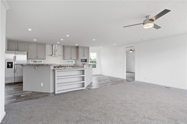 kitchen featuring ceiling fan, an island with sink, wall chimney exhaust hood, gray cabinetry, and appliances with stainless steel finishes