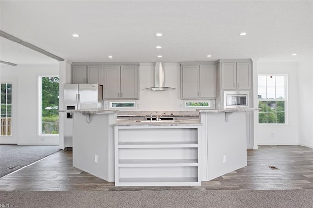 kitchen with appliances with stainless steel finishes, wall chimney range hood, a kitchen island with sink, and dark wood-type flooring