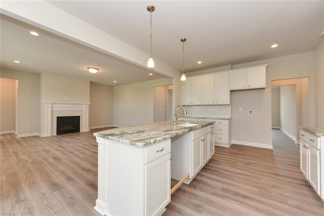 kitchen with light stone counters, light hardwood / wood-style floors, hanging light fixtures, a center island with sink, and white cabinets