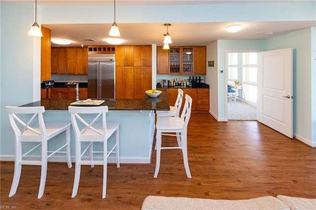 kitchen with dark stone counters, hardwood / wood-style floors, stainless steel built in refrigerator, hanging light fixtures, and a breakfast bar