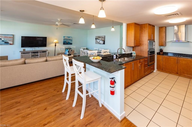 kitchen with dark stone counters, kitchen peninsula, decorative light fixtures, wall chimney exhaust hood, and sink