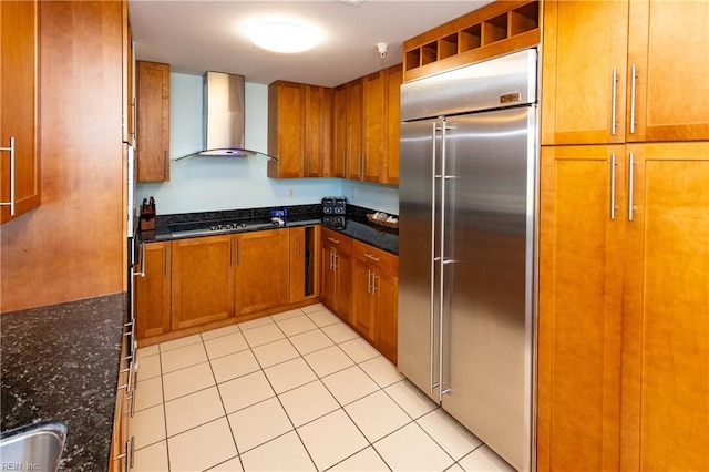 kitchen with black gas cooktop, built in fridge, wall chimney exhaust hood, dark stone countertops, and light tile floors