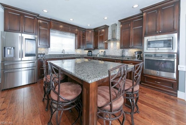 kitchen featuring stainless steel appliances, dark wood-type flooring, a breakfast bar, a kitchen island, and wall chimney range hood