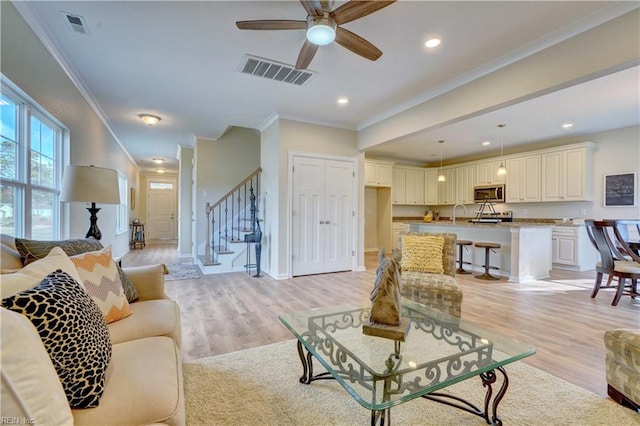 living area with stairs, light wood-style floors, visible vents, and ornamental molding