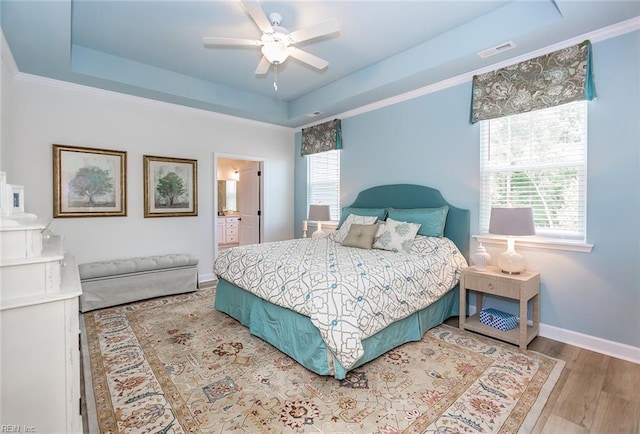 bedroom featuring ornamental molding, light wood-type flooring, ensuite bathroom, a tray ceiling, and ceiling fan