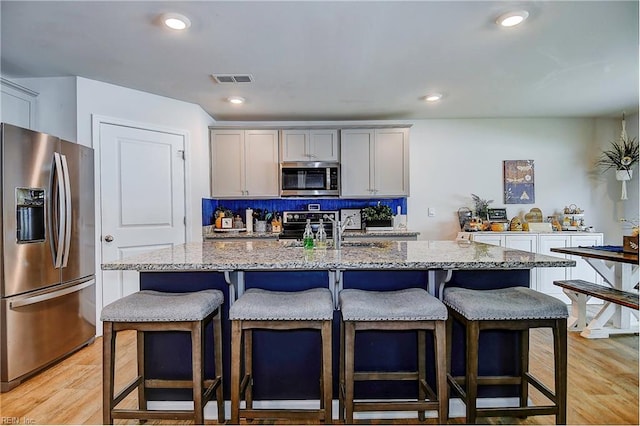 kitchen with appliances with stainless steel finishes, backsplash, light stone counters, an island with sink, and light wood-type flooring