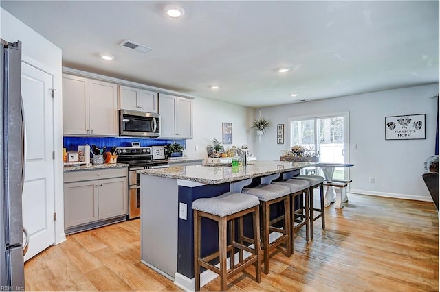 kitchen featuring appliances with stainless steel finishes, decorative backsplash, light stone counters, light hardwood / wood-style floors, and a center island with sink