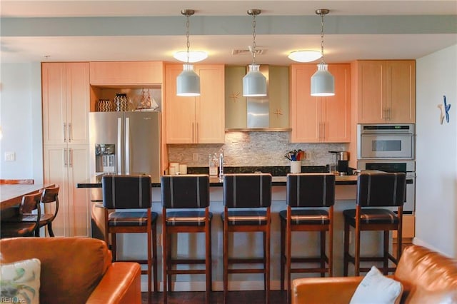 kitchen featuring decorative light fixtures, a kitchen breakfast bar, decorative backsplash, light brown cabinets, and wall chimney range hood