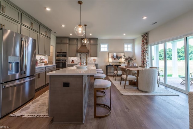 kitchen featuring wood-type flooring, a kitchen island with sink, backsplash, appliances with stainless steel finishes, and decorative light fixtures