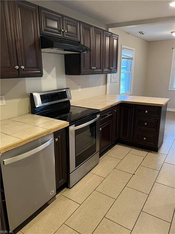 kitchen with light tile patterned flooring, kitchen peninsula, dark brown cabinets, appliances with stainless steel finishes, and tile counters