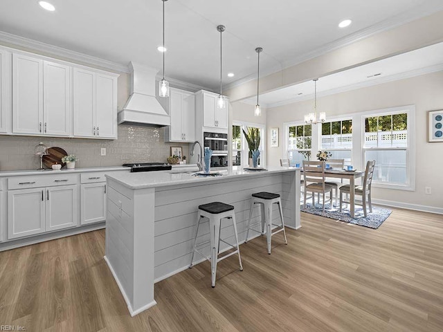 kitchen featuring a kitchen island with sink, white cabinets, premium range hood, light hardwood / wood-style flooring, and decorative light fixtures