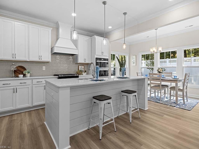 kitchen with white cabinets, an island with sink, hanging light fixtures, custom range hood, and light hardwood / wood-style floors