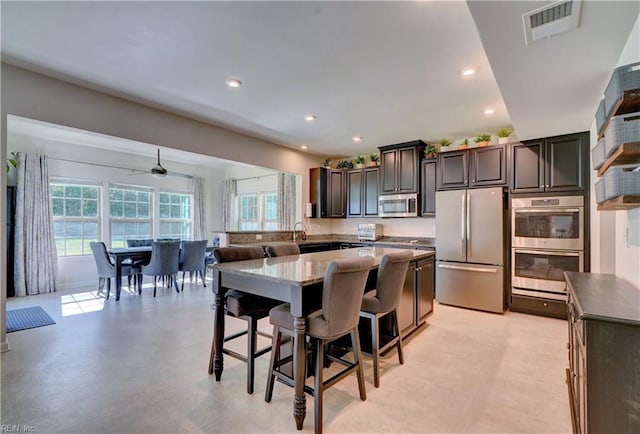 kitchen featuring ceiling fan, a kitchen island, dark brown cabinets, appliances with stainless steel finishes, and a kitchen breakfast bar