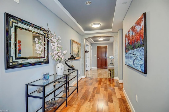 hall featuring light wood-style flooring, a tray ceiling, baseboards, and arched walkways