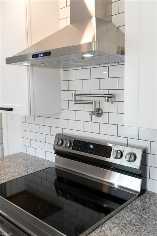 kitchen featuring backsplash, light stone counters, stainless steel electric stove, wall chimney range hood, and white cabinets