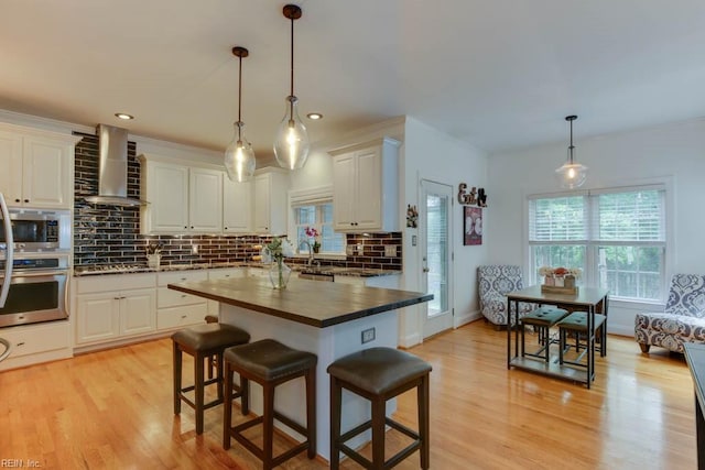 kitchen with stainless steel appliances, light hardwood / wood-style floors, wall chimney exhaust hood, decorative light fixtures, and a center island