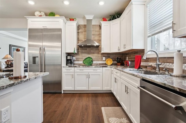 kitchen featuring wall chimney range hood, sink, white cabinetry, appliances with stainless steel finishes, and light stone counters