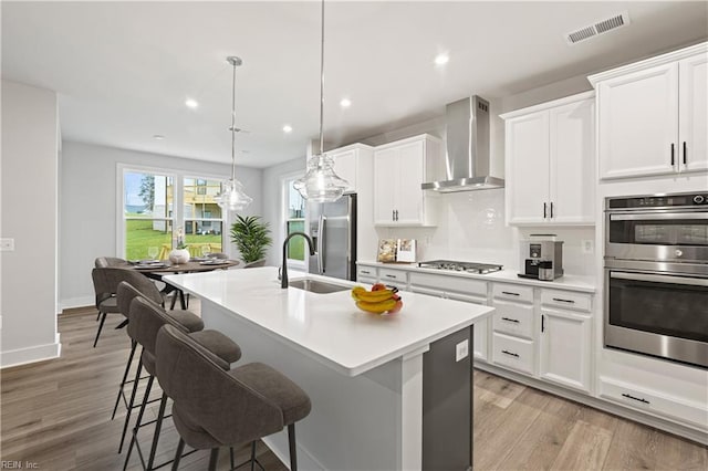 kitchen featuring wall chimney range hood, an island with sink, hanging light fixtures, white cabinets, and appliances with stainless steel finishes