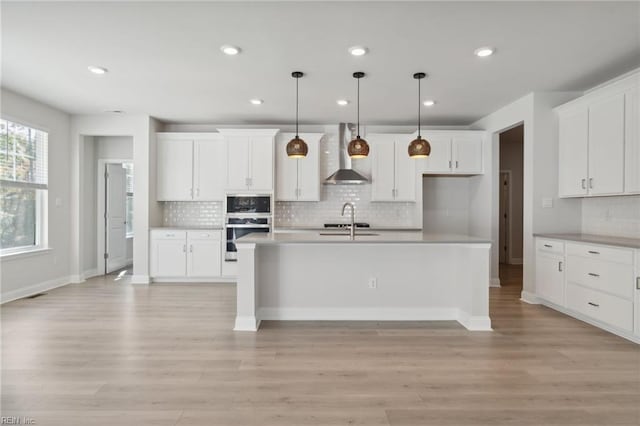 kitchen with a kitchen island with sink, wall chimney range hood, light hardwood / wood-style flooring, decorative light fixtures, and white cabinetry