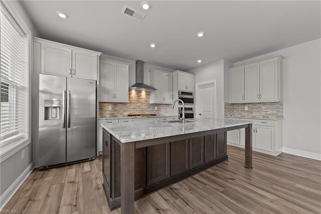 kitchen featuring a kitchen island with sink, appliances with stainless steel finishes, light hardwood / wood-style floors, and wall chimney range hood