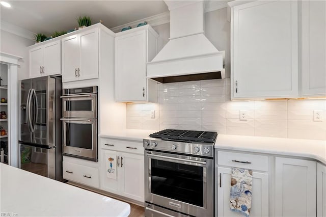 kitchen featuring white cabinetry, appliances with stainless steel finishes, decorative backsplash, custom range hood, and ornamental molding