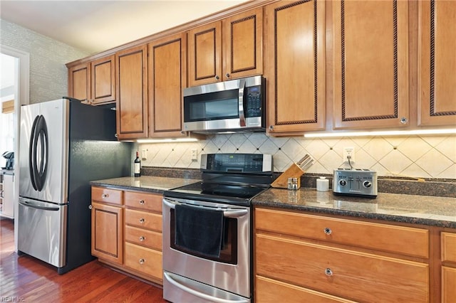 kitchen with dark stone counters, stainless steel appliances, brown cabinetry, and dark wood-style flooring