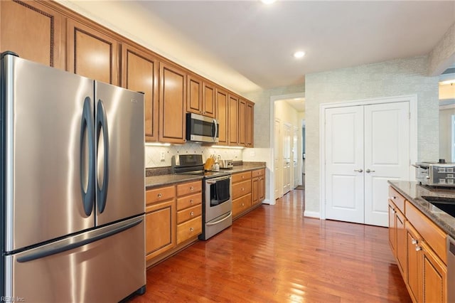 kitchen featuring brown cabinets, stainless steel appliances, backsplash, dark wood-type flooring, and dark stone counters