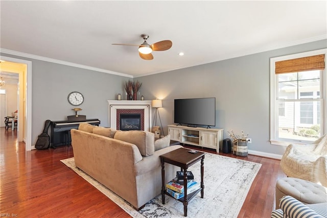 living room featuring a tiled fireplace, dark wood-type flooring, ornamental molding, ceiling fan, and baseboards