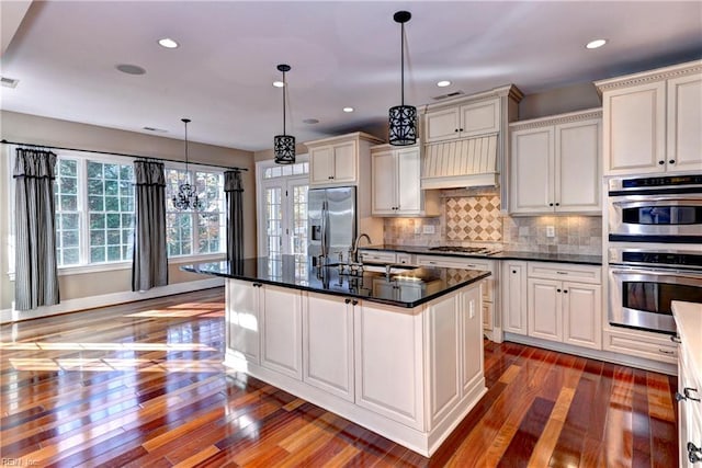 kitchen featuring backsplash, a center island with sink, hanging light fixtures, appliances with stainless steel finishes, and dark hardwood / wood-style flooring