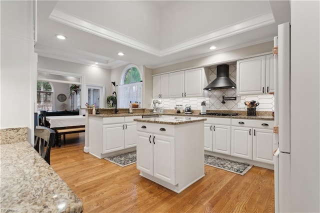 kitchen featuring a center island, wall chimney range hood, light hardwood / wood-style flooring, crown molding, and white cabinets