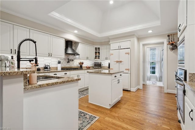 kitchen with a tray ceiling, white cabinets, a kitchen island, and wall chimney range hood