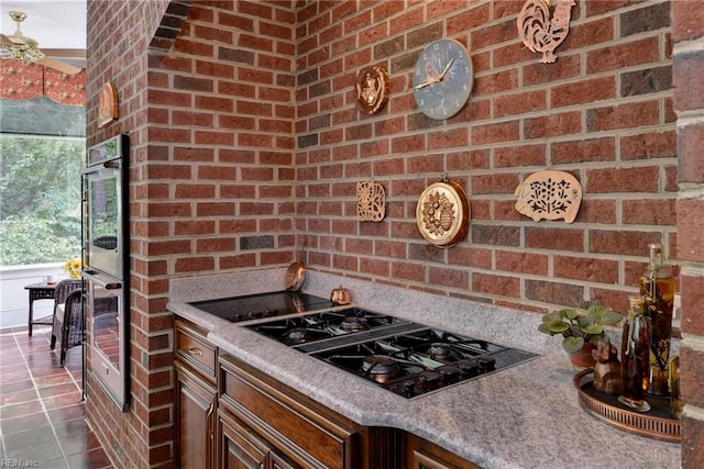 kitchen featuring black gas stovetop, stainless steel double oven, light stone counters, and brick wall