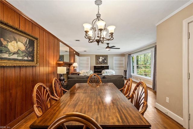 dining space with ornamental molding, ceiling fan with notable chandelier, wooden walls, and dark wood-type flooring