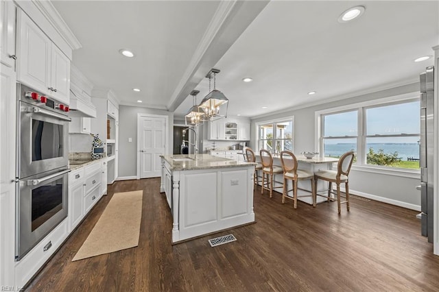 kitchen featuring double oven, white cabinets, a water view, and an island with sink