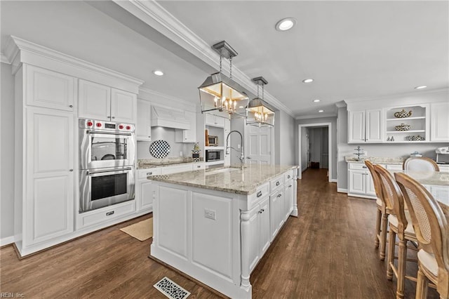 kitchen with dark wood-type flooring, hanging light fixtures, stainless steel double oven, a kitchen island with sink, and white cabinets