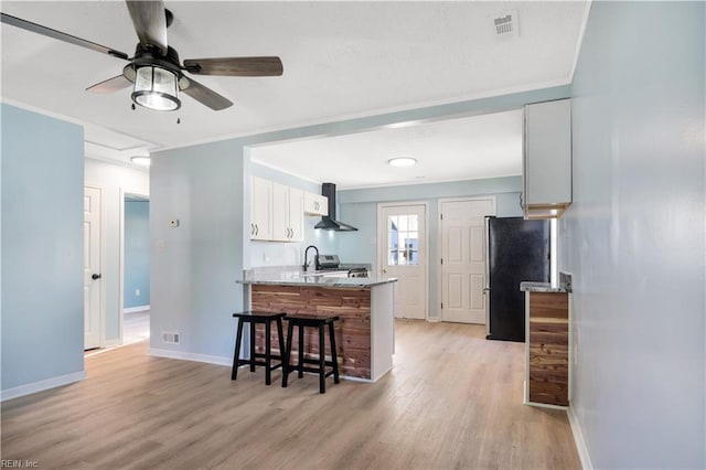 kitchen with white cabinetry, wall chimney exhaust hood, light hardwood / wood-style flooring, kitchen peninsula, and black refrigerator