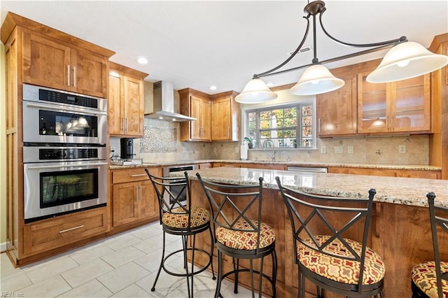 kitchen with tasteful backsplash, light stone counters, wall chimney exhaust hood, stainless steel appliances, and decorative light fixtures