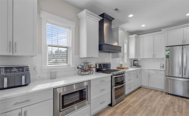 kitchen with light stone counters, wall chimney exhaust hood, stainless steel appliances, white cabinets, and light hardwood / wood-style floors