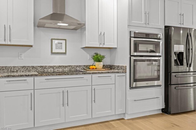 kitchen with stainless steel appliances, light hardwood / wood-style floors, wall chimney exhaust hood, and dark stone countertops