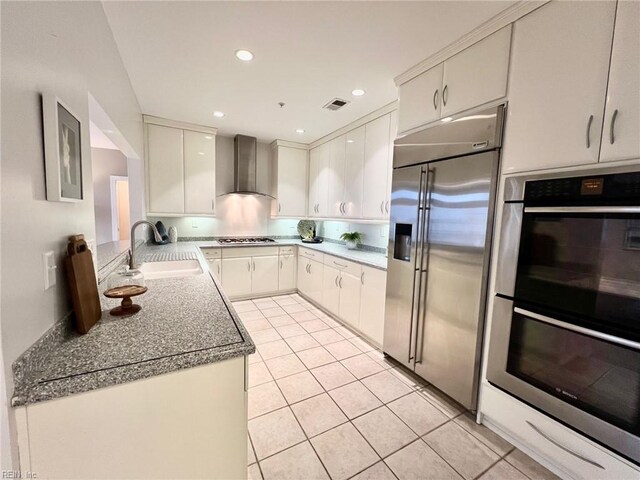 kitchen featuring wall chimney range hood, sink, light tile patterned floors, appliances with stainless steel finishes, and white cabinetry