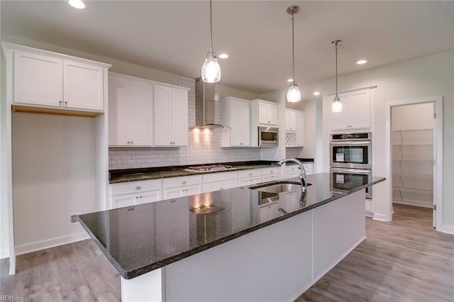 kitchen with sink, wall chimney exhaust hood, an island with sink, white cabinets, and appliances with stainless steel finishes