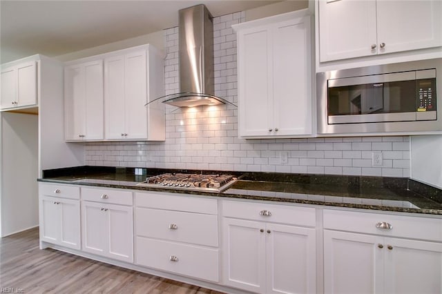 kitchen featuring stainless steel appliances, white cabinetry, dark stone counters, and wall chimney range hood