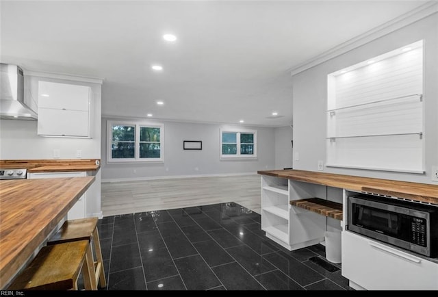 kitchen with stainless steel microwave, wall chimney exhaust hood, wooden counters, white cabinets, and ornamental molding