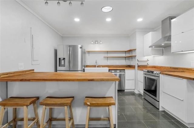 kitchen featuring sink, stainless steel appliances, wall chimney range hood, butcher block countertops, and a breakfast bar area