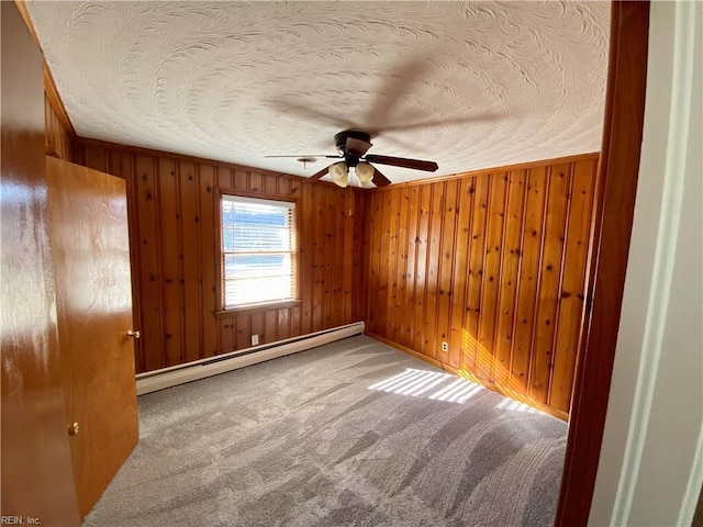 carpeted empty room featuring a textured ceiling, ceiling fan, a baseboard heating unit, and wood walls